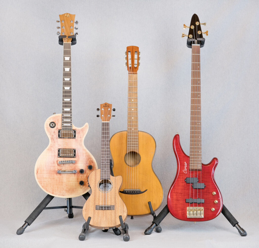 Ansicht aufgestellter Gitarren in Ständern. Von links nach rechts: E-Gitarre, Ukulele, klassische Gitarre und E-Bass-Gitarre